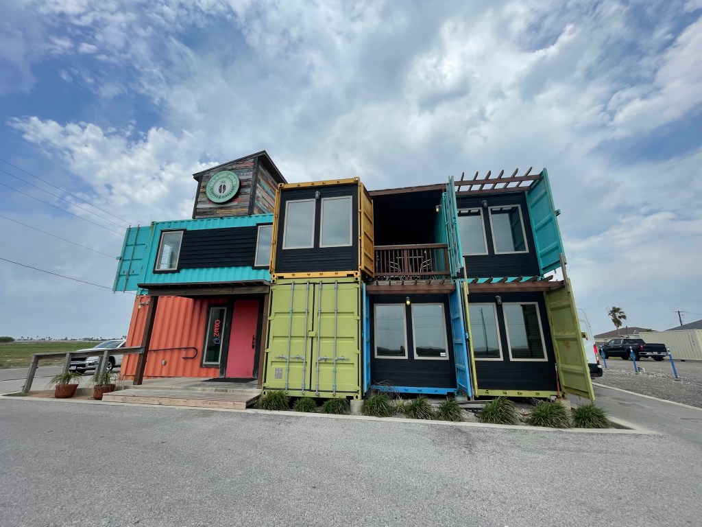 Exterior view of a modern building made from colorful shipping containers, with a clock on top and a wooden deck entrance, set against a cloudy sky.
